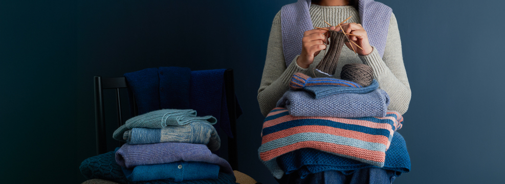 A woman knitting a sock, surrounded by layered hand-knit sweaters, shawls, hats, and blankets, modeling knit-for-yourself knitting patterns.